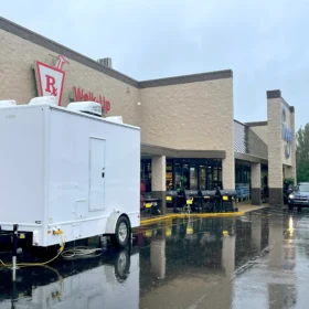 A white restroom trailer is stationed outside a Kroger grocery store near the walk-up pharmacy entrance on a rainy day, with wet pavement reflecting the building and vehicles.