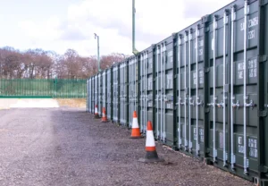 A neat row of dark green 40-foot storage containers lines a fenced gravel yard, with orange traffic cones placed along the path under a cloudy sky.