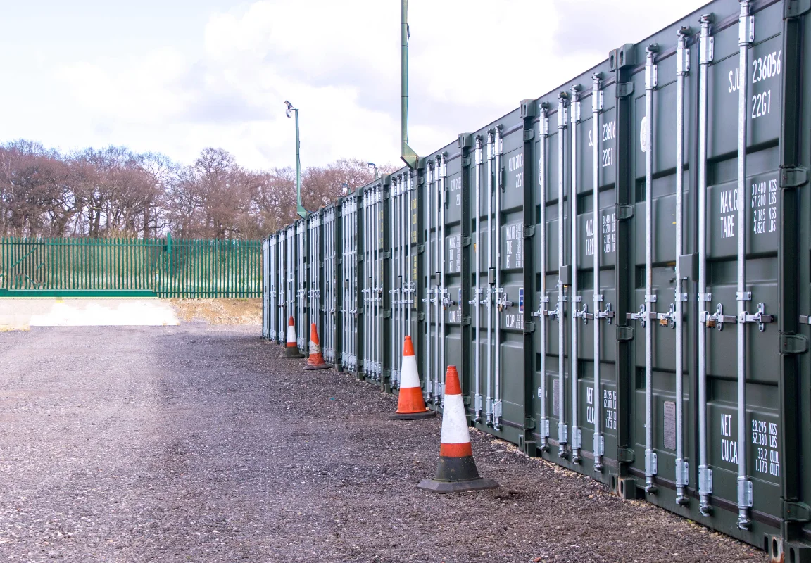 A neat row of dark green 40-foot storage containers lines a fenced gravel yard, with orange traffic cones placed along the path under a cloudy sky.