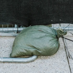 A close-up of a green sandbag tied around the base of a temporary fence frame, providing extra stability for the privacy screen.