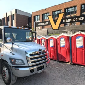 A white sanitation truck is parked on gravel next to a row of red portable restrooms, with a large Vanderbilt Commodores sign visible on a brick building in the background.