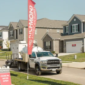A white sanitation truck with a silver tank is parked on a residential street beside two red flags and a sign for Richmond American Homes, with new houses visible in the background.