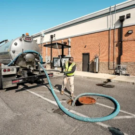 A sanitation truck with a shiny metal tank is parked in a lot beside a brick building, where a worker in a safety vest is using a hose to service a manhole.
