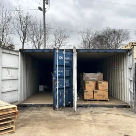 An open shipping container filled with stacked cardboard boxes and packing materials is positioned on a gravel surface beside a portable restroom, with bare trees and overcast skies in the background.