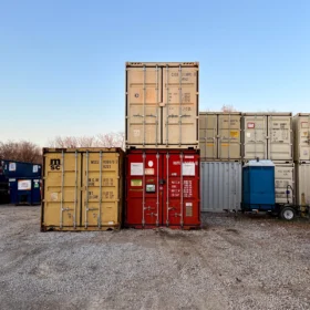 A stack of shipping containers in various colors is positioned in a gravel lot beside a white delivery truck and a blue portable restroom, under a clear blue sky.