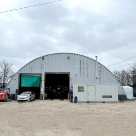 A silver car is parked in front of a large metal warehouse with a green tarp covering the entrance, alongside a red skid steer loader and a white water tank on a cloudy day.