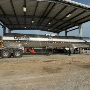 A large silver sanitation truck with the "FusionSite" logo is parked under a metal shelter on a gravel surface, with orange safety cones and other vehicles visible in the background.