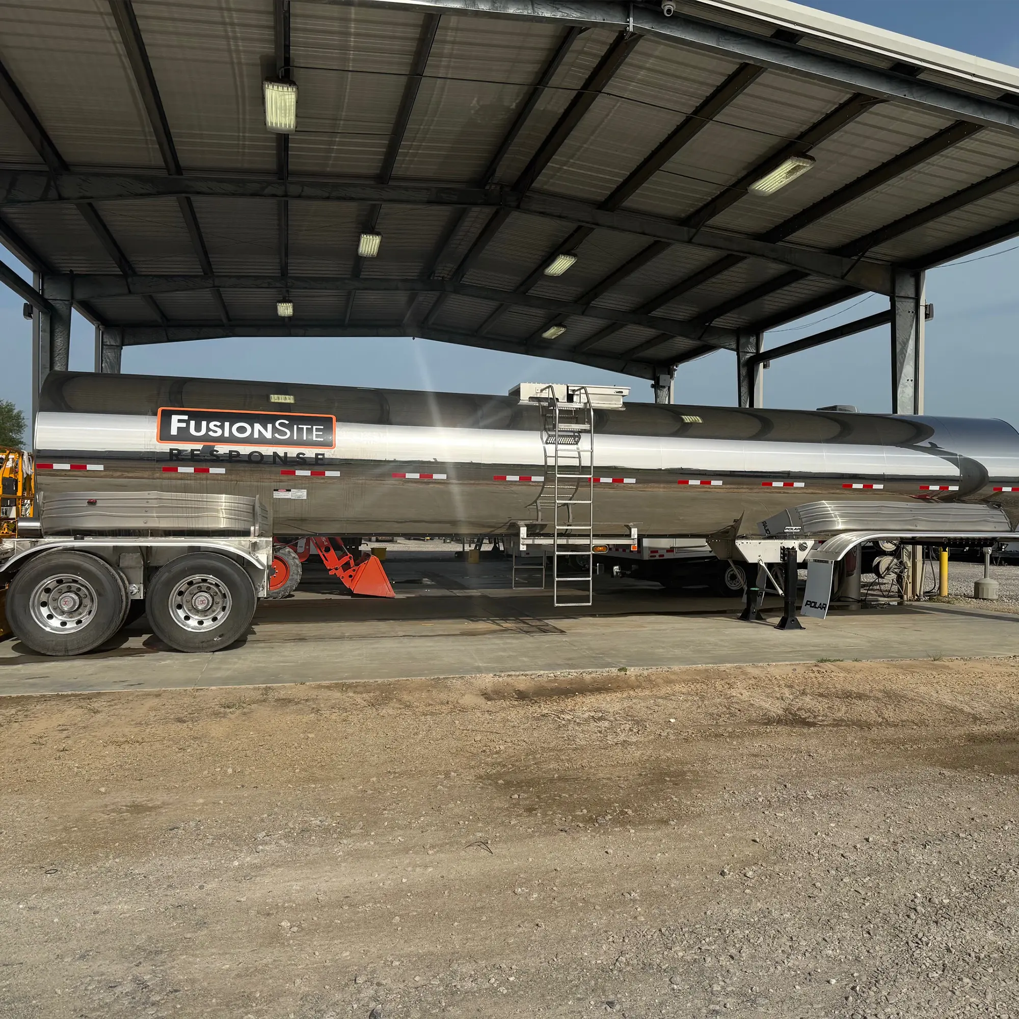 A large silver sanitation truck with the "FusionSite" logo is parked under a metal shelter on a gravel surface, with orange safety cones and other vehicles visible in the background.