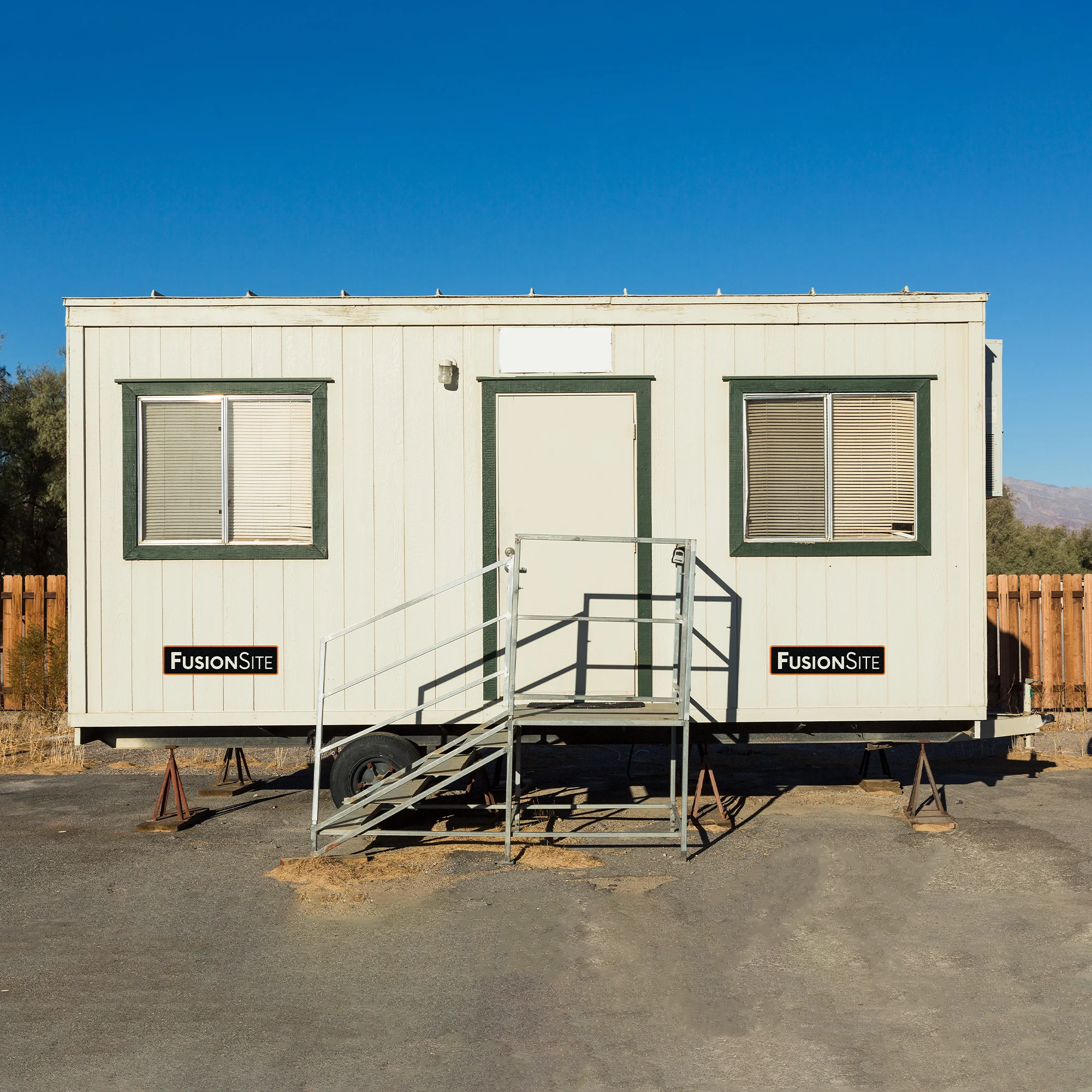 A white portable office trailer with two windows and a door is positioned on a gravel lot, next to a wooden fence and surrounded by sparse vegetation under a clear blue sky.