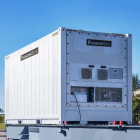 A white portable refrigerated unit with ventilation and branding is positioned on a gravel lot, surrounded by trees and a clear blue sky in the background.