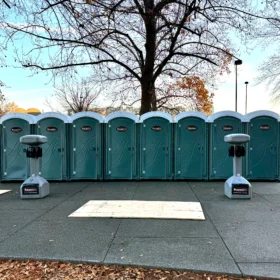 A row of teal portable restrooms is positioned on a paved area beneath bare trees, with handwashing stations nearby and a fenced construction site in the background on a clear day.