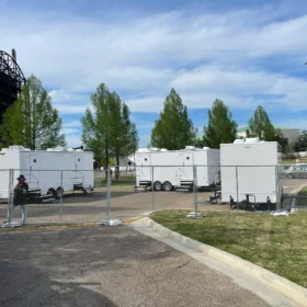 White restroom trailers are enclosed by chain-link temporary fencing at an outdoor event under a bright sky.