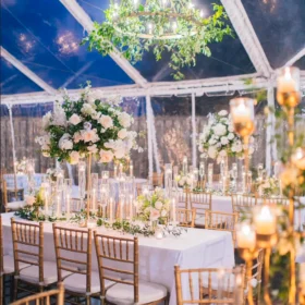 A beautifully decorated tented event space features elegantly set tables with floral centerpieces, tall glass candle holders, and golden chairs, all under a blue canopy with twinkling lights.