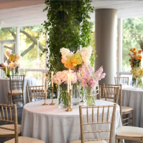 A beautifully decorated wedding reception table with gold chairs, pastel floral centerpieces, and tall candles surrounded by greenery and large windows letting in natural light.