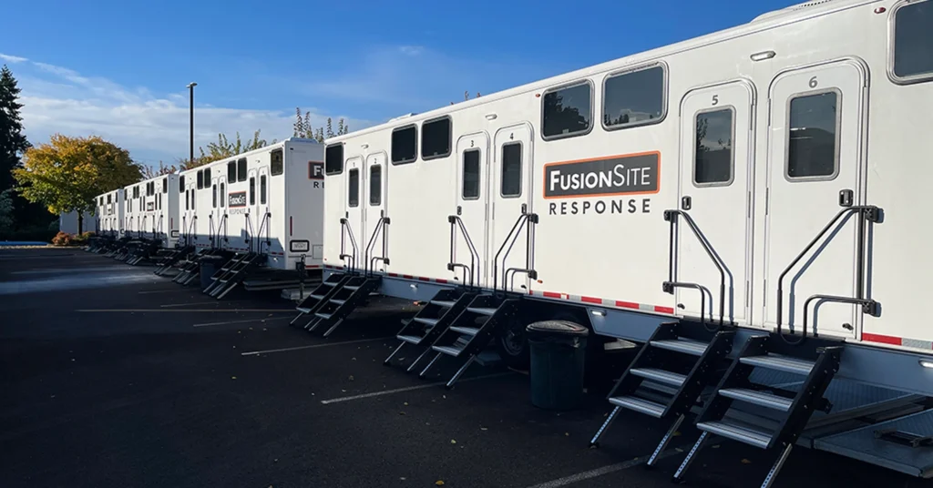 A row of white portable restroom trailers with steps is parked in a paved lot, surrounded by trees and under a clear blue sky.