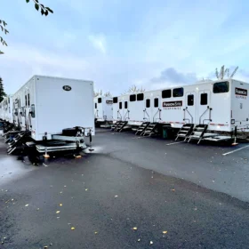 A row of white portable restroom trailers with steps is parked on a paved lot, surrounded by trees and under a cloudy sky.