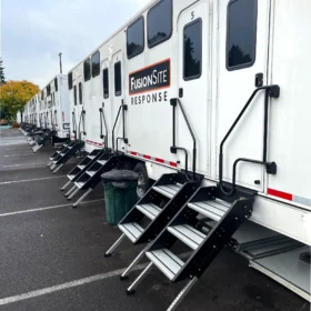 A row of white portable restroom trailers with steps is parked in a paved lot, surrounded by trees and under a cloudy sky.