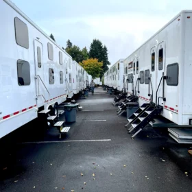 A row of white portable restroom trailers is positioned in a parking lot, flanked by trash bins and surrounded by trees under a cloudy sky.