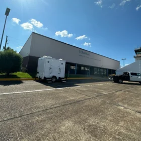 A white restroom trailer is parked outside the Hawkins Field Airport terminal in Jackson, Mississippi, under a bright blue sky with scattered clouds.