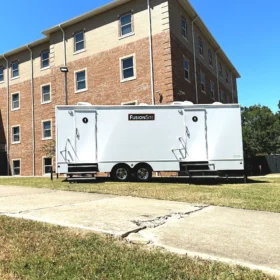 A white FusionSite restroom trailer with dual entrances is parked beside a brick campus building on a sunny day with a clear blue sky.