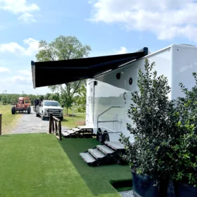 A white restroom trailer with extended black awning and metal stairs is set up on a grassy field alongside parked trucks under a bright blue sky.