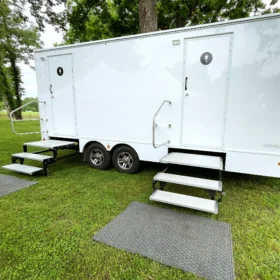 A white restroom trailer with dual entry doors, handrails, and steps is stationed on a grassy lawn surrounded by trees and event tents.