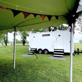 A white FusionSite restroom trailer with twin doors and stairs is set up on green grass beneath an outdoor event tent with hanging banners.