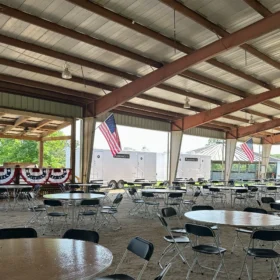 Two white FusionSite restroom trailers are parked outside a large covered pavilion decorated with American flags and event tables.