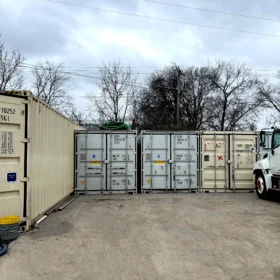 A white service truck is parked beside a row of metal shipping containers in a gravel lot, with bare trees and a cloudy sky in the background.