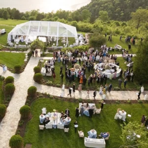 A large white tent is set up on a grassy hillside, with guests mingling and seated at tables, while a scenic view of the water and trees is visible in the background.
