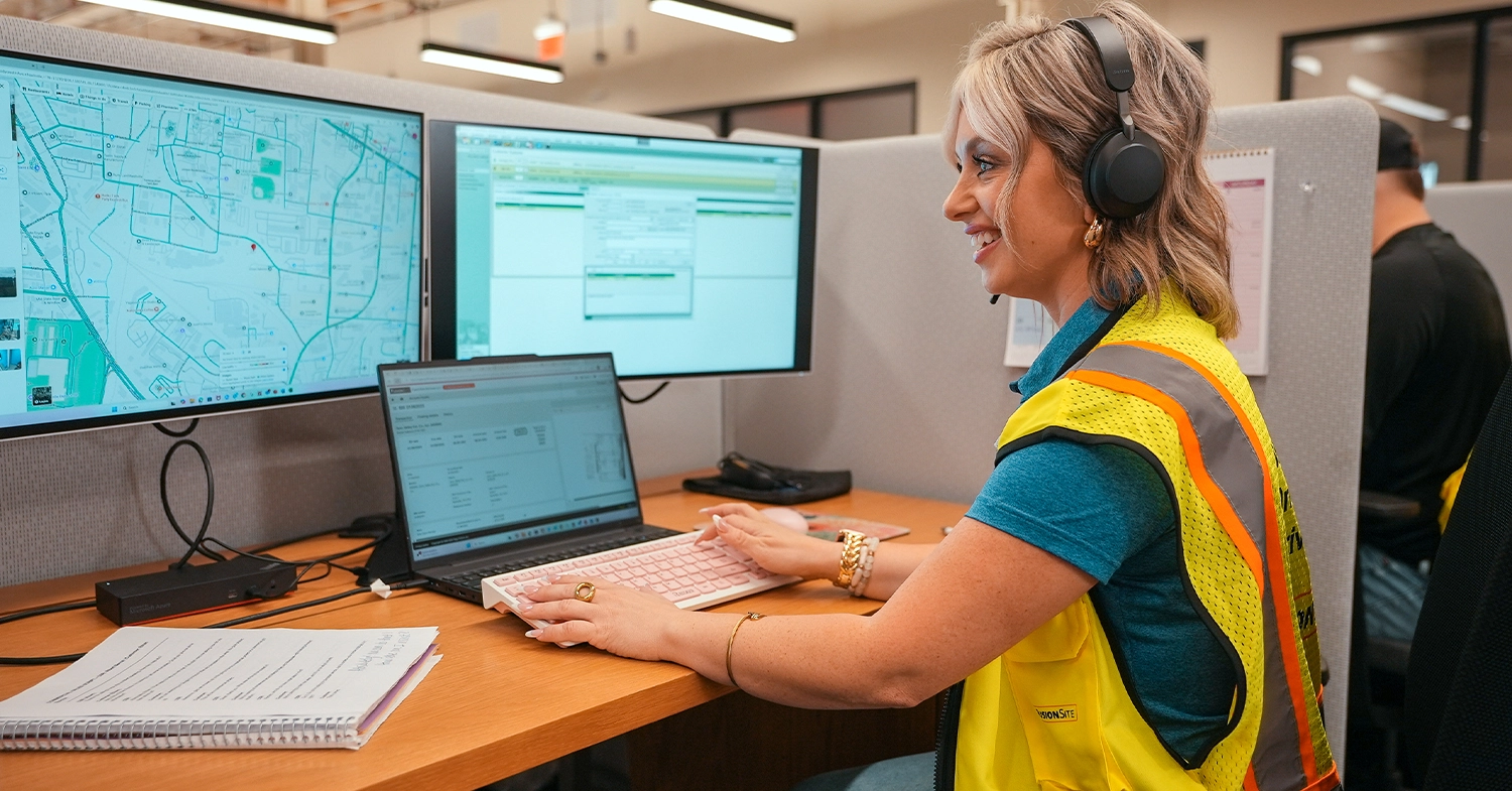 A woman wearing a yellow safety vest and headphones is seated at a desk, working on a laptop and two monitors displaying maps and data in a brightly lit office environment.