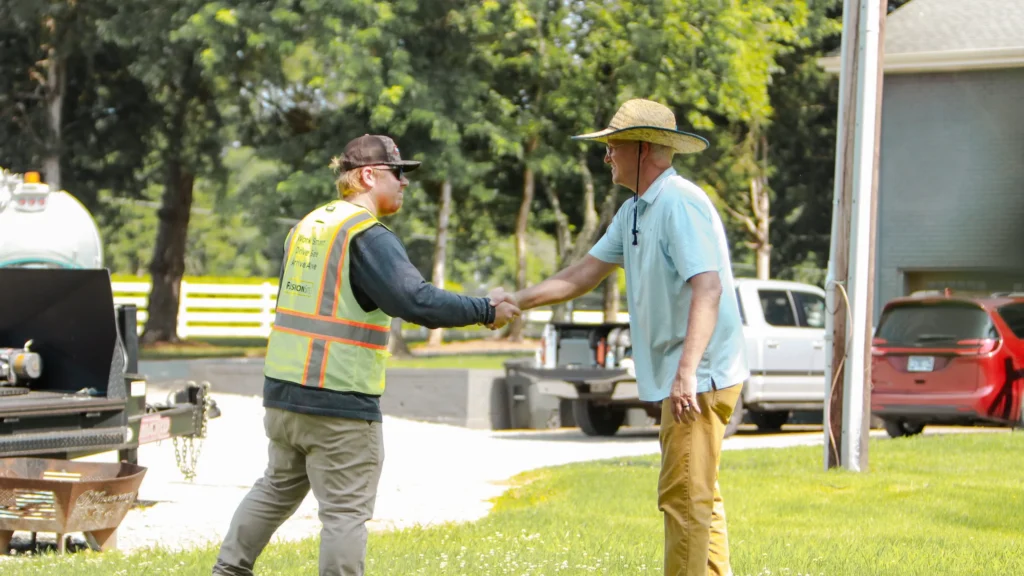 A worker in a reflective vest shakes hands with a man in a blue shirt on a grassy area beside a parked truck and a white fence, surrounded by trees on a sunny day.