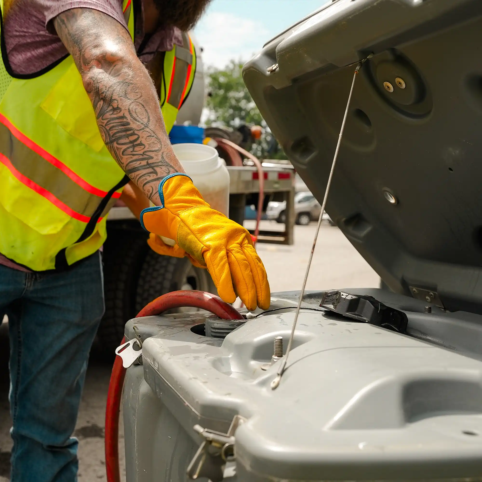 A worker in a yellow safety vest and gloves is inspecting the open lid of a portable restroom tank while standing in a parking lot with vehicles in the background on a sunny day.