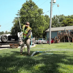A worker in a reflective vest uses a long hose near a parked truck and a two-story building, surrounded by grass and trees on a sunny day.