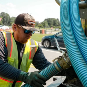 A worker in a reflective vest and gloves connects a blue hose to a sanitation truck while parked on a paved area, with vehicles and green grass visible in the background on a sunny day.