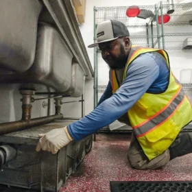 A worker in a yellow safety vest and gloves kneels beside a metal sink, inspecting a drainage system in a clean, well-lit service area with red flooring.