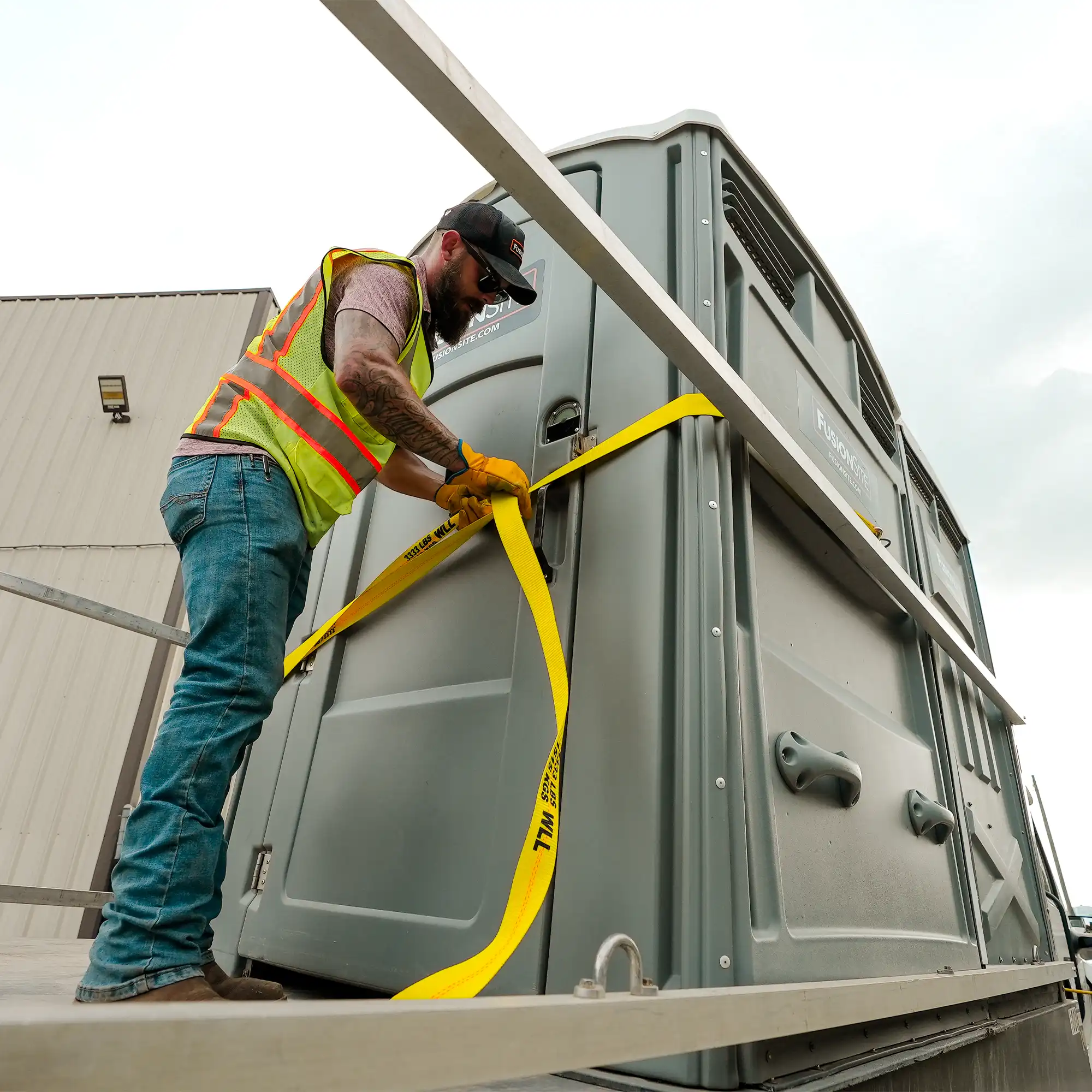 A worker in a safety vest secures a portable restroom with yellow straps on a delivery truck beside a large warehouse, under a cloudy sky.