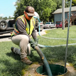 A worker in a reflective vest and gloves kneels on grass, connecting a blue hose to a green septic tank access point, with parked vehicles and a wooden deck in the background on a sunny day.