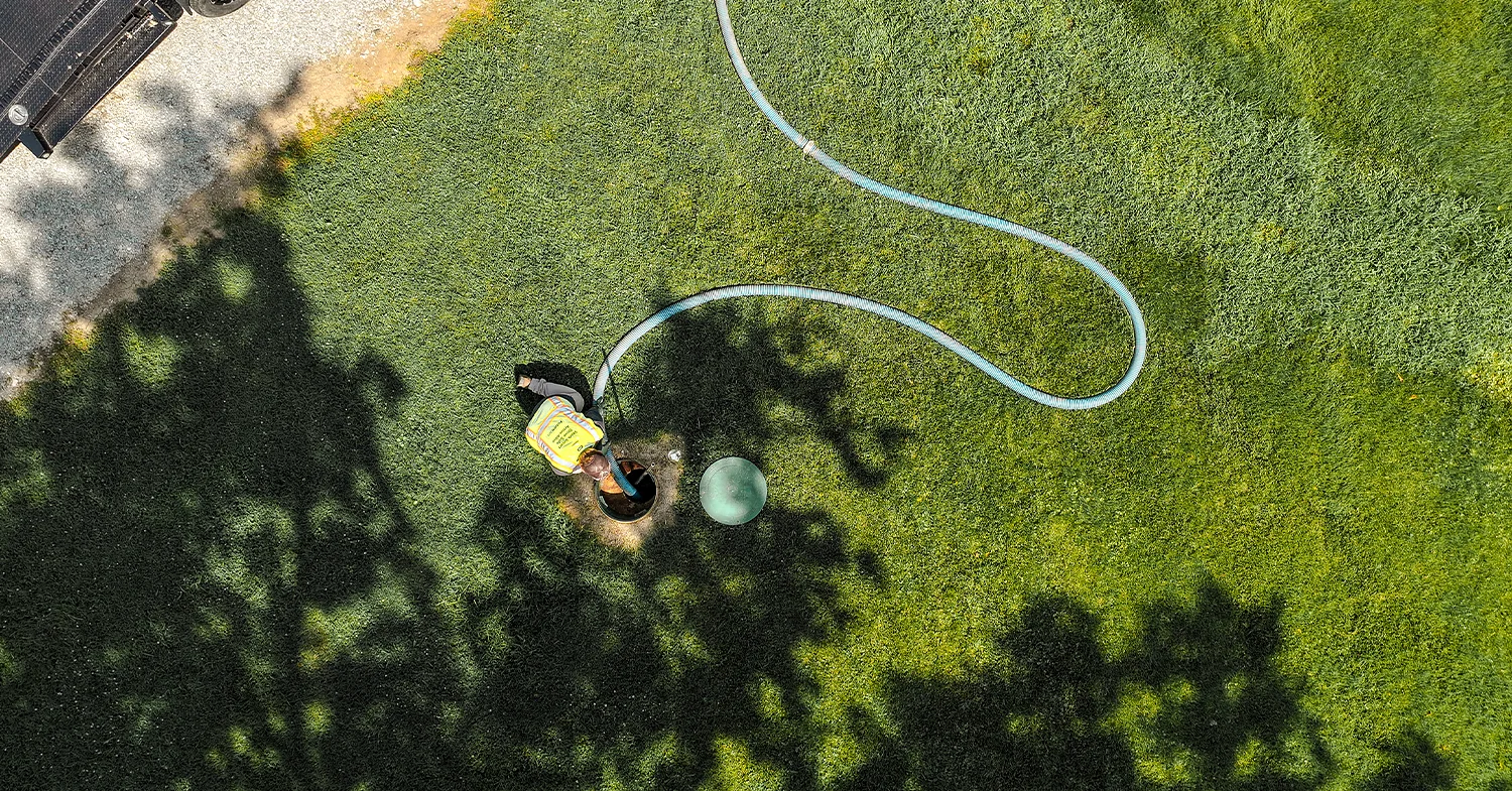 A worker in a safety vest is using a vacuum hose to service a green septic tank on a grassy area, with a gravel path visible in the background.