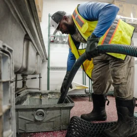 A worker in a yellow safety vest and gloves is using a hose to pump waste from a gray tank located in a clean, indoor service area with metal shelving and a red rubber floor mat.