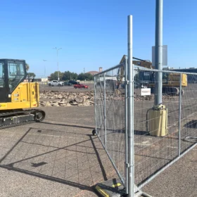 A yellow mini excavator is parked on a gravel lot beside a construction site, enclosed by a chain-link fence, with a clear blue sky overhead and distant vehicles visible in the background.